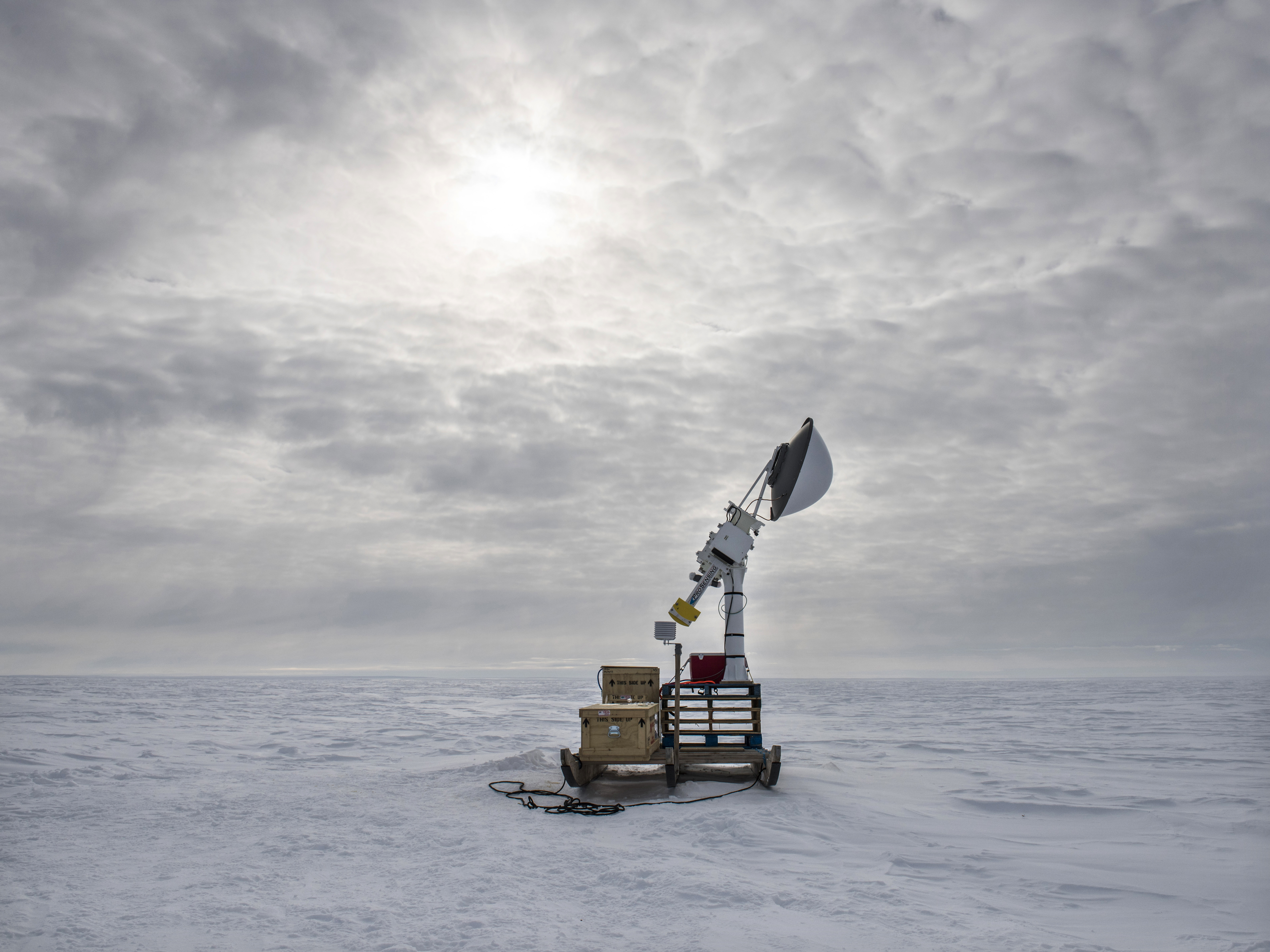 Two qamutiks beside each other on ice with a cloudy sky in the background. Two wooden boxes are stacked on the left qamutik. Pallets are stacked on the right qamutik with a large antenna structure on top.